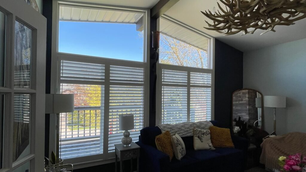 A modern, open-concept living room featuring a row of three glass patio doors fitted with custom bifold plantation shutters. The white-painted wood shutters are partially folded open to reveal the window panes and the outdoor patio deck beyond. When opened, the stacked shutter panels neatly frame each glass section, coordinating seamlessly with the light hardwood flooring and clean interior styling.
