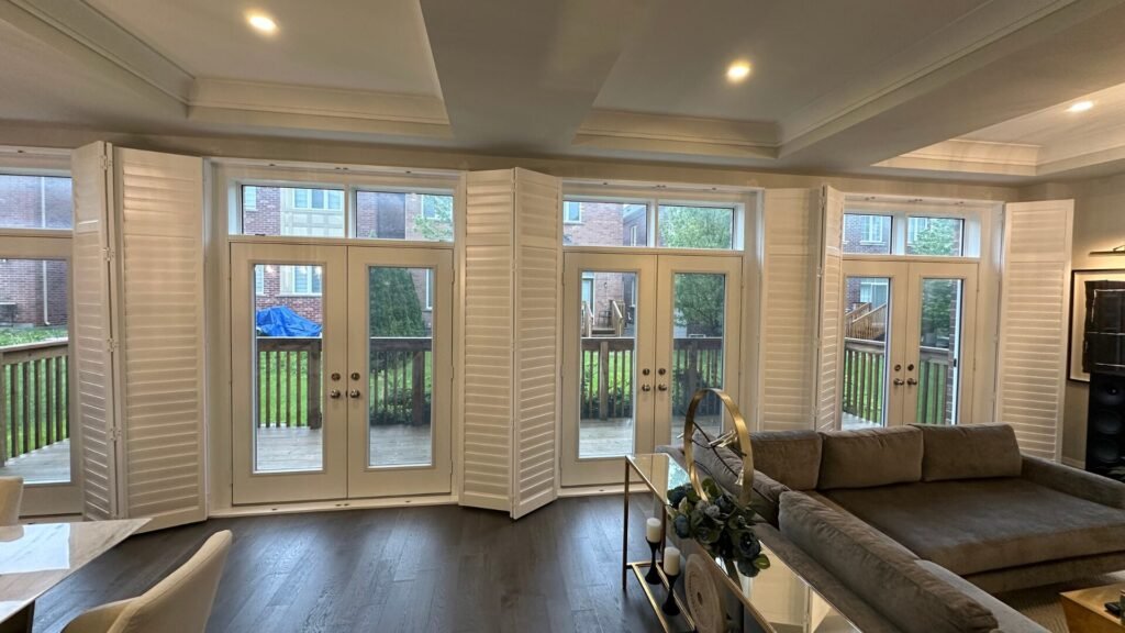 A wide living room featuring three sets of double glass patio doors leading to a deck. Each door assembly is framed with white, multi-panel plantation shutters (bifold style) that are partially folded open to reveal the glass panels.