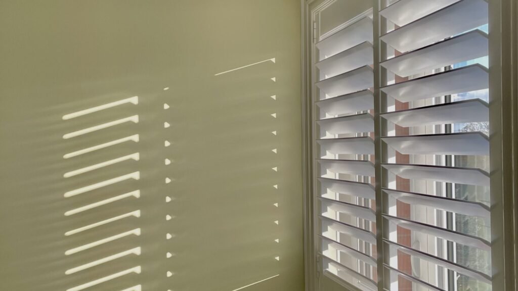 A close-up view of premium window shutters installed in a Toronto home, highlighting the fine craftsmanship and clean design. The image showcases the sleek white louvers of the custom plantation shutters, precisely aligned to control light and privacy. The crisp white finish of the shutter frame and slats stands out against a neutral wall backdrop, illustrating a popular, modern window treatment style found throughout the Greater Toronto Area (GTA).