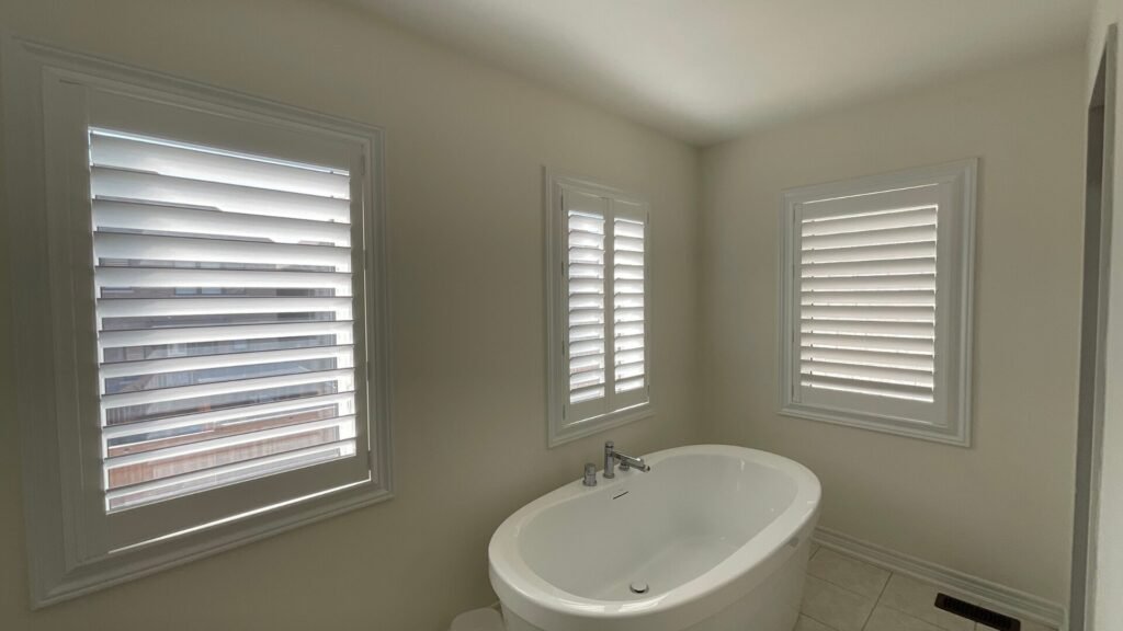 A bright, modern bathroom featuring a window fitted with custom white plantation shutters. The bifold shutter panels are folded completely open to the left side, exposing the window pane and allowing natural daylight to stream into the space. A portion of a glass shower enclosure or vanity mirror is visible on the right, reflecting the clean white walls of the room.