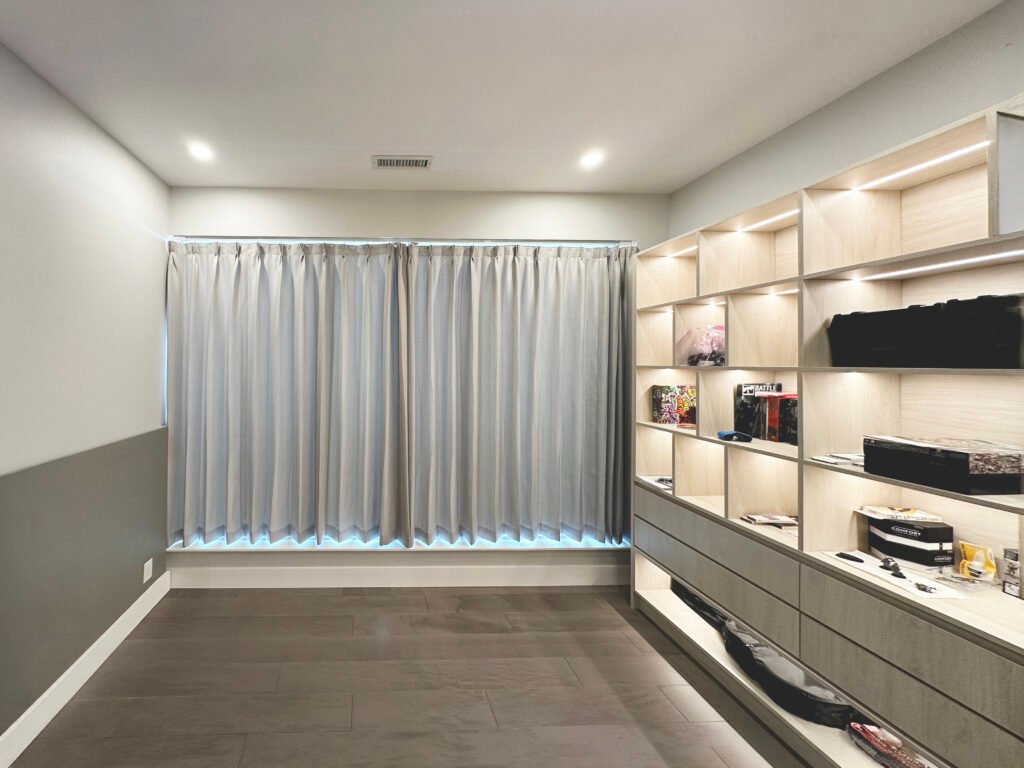 A sleek, modern room featuring a wide window covered by light grey, pinch-pleat blackout curtains. The drapes run along a ceiling-mounted track and are pulled completely shut, hanging just above a low window ledge. Subtle daylight peeks out from the very top and bottom edges of the curtains, creating a soft halo effect. To the right, a large, built-in light wood shelving unit stretches across the wall, illuminated by warm, integrated LED strip lighting on each shelf. The room is finished with grey walls, white baseboards, and dark grey stone-look floor tiles.