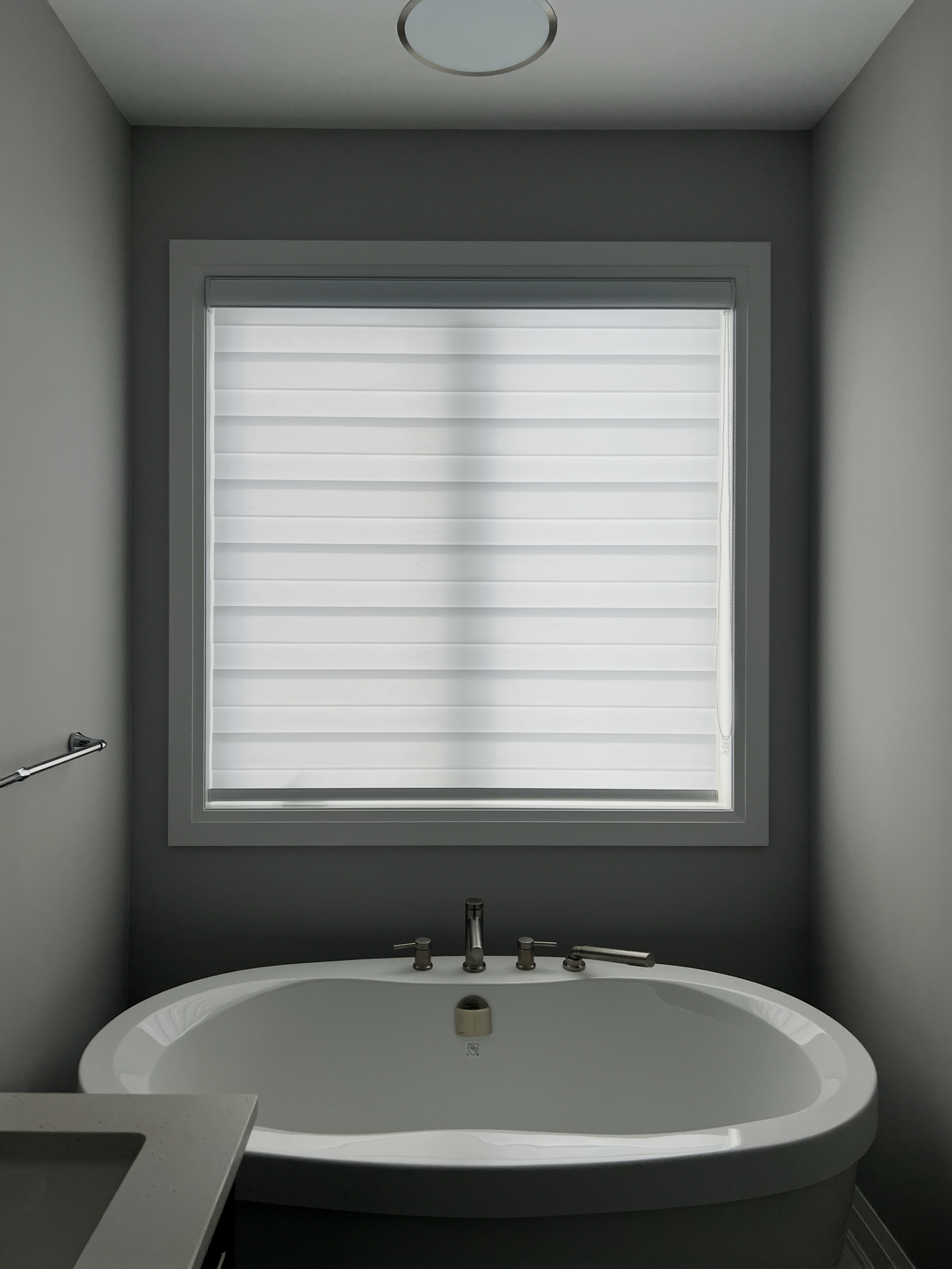 A sleek white zebra blind covering a square window directly above an oval-shaped freestanding bathtub in a moody gray tiled bathroom.