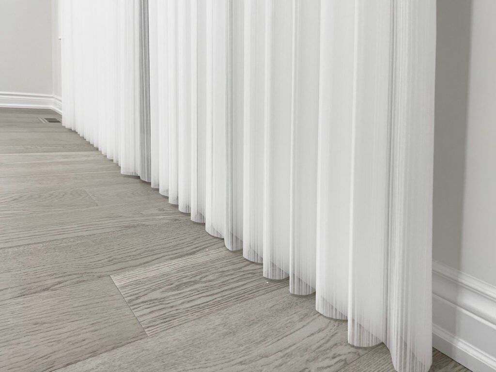 Detailed view of flowing white sheer dream shades resting elegantly on a grey hardwood floor, combining the soft look of a curtain with the utility of a blind.