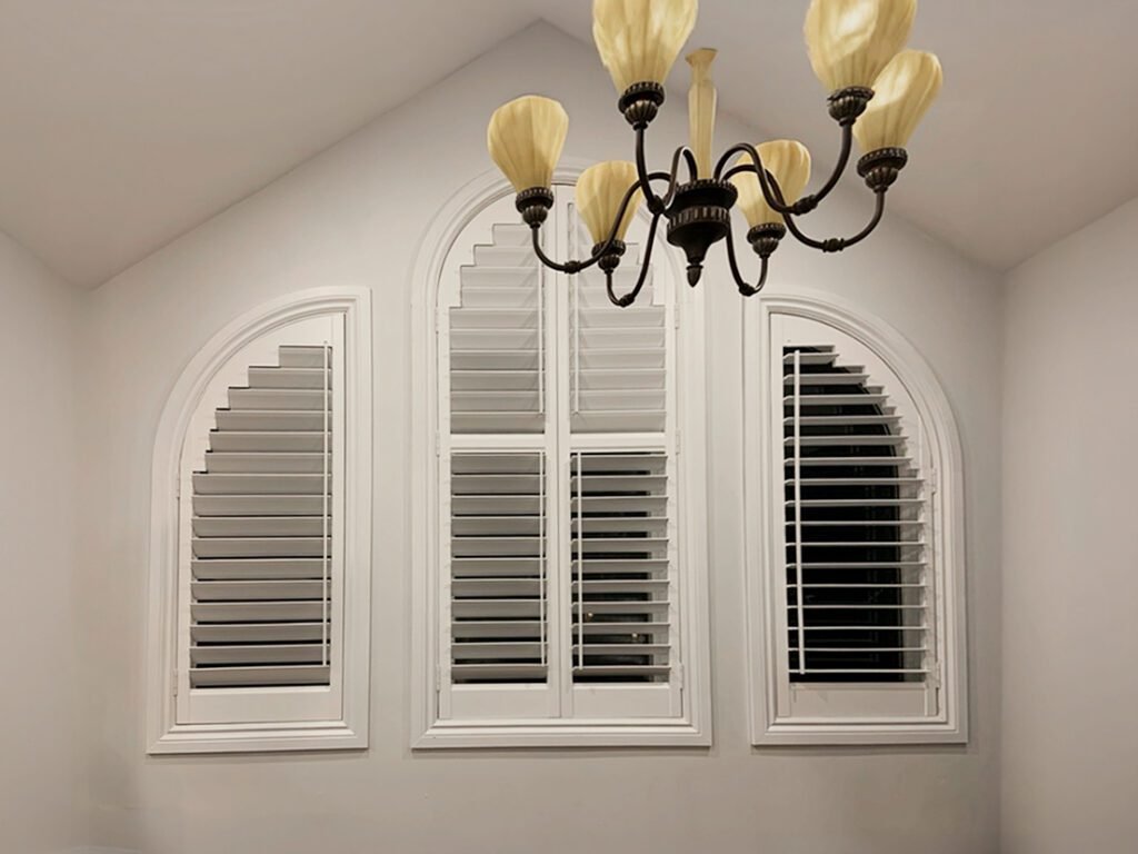 Classic white plantation shutters custom-fitted for three arched architectural windows beneath a decorative chandelier.