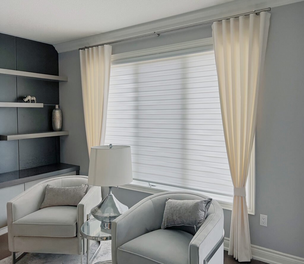 A modern grey sitting room displaying a window treated with a white zebra blind and layered with decorative cream-colored curtain side panels on a silver rod.
