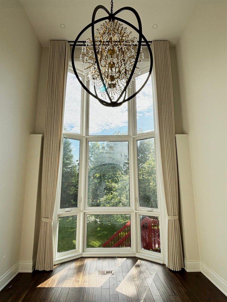 Tall beige drapery panels framed perfectly around a massive two-story bay window overlooking a backyard, situated beneath a crystal orb chandelier.
