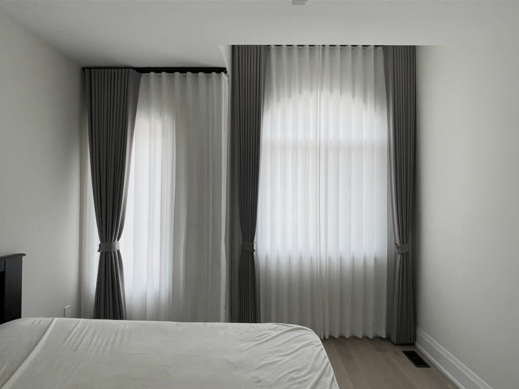 Custom floor-to-ceiling bedroom drapery in gray paired with white sheer curtains, neatly tied back to frame the windows.