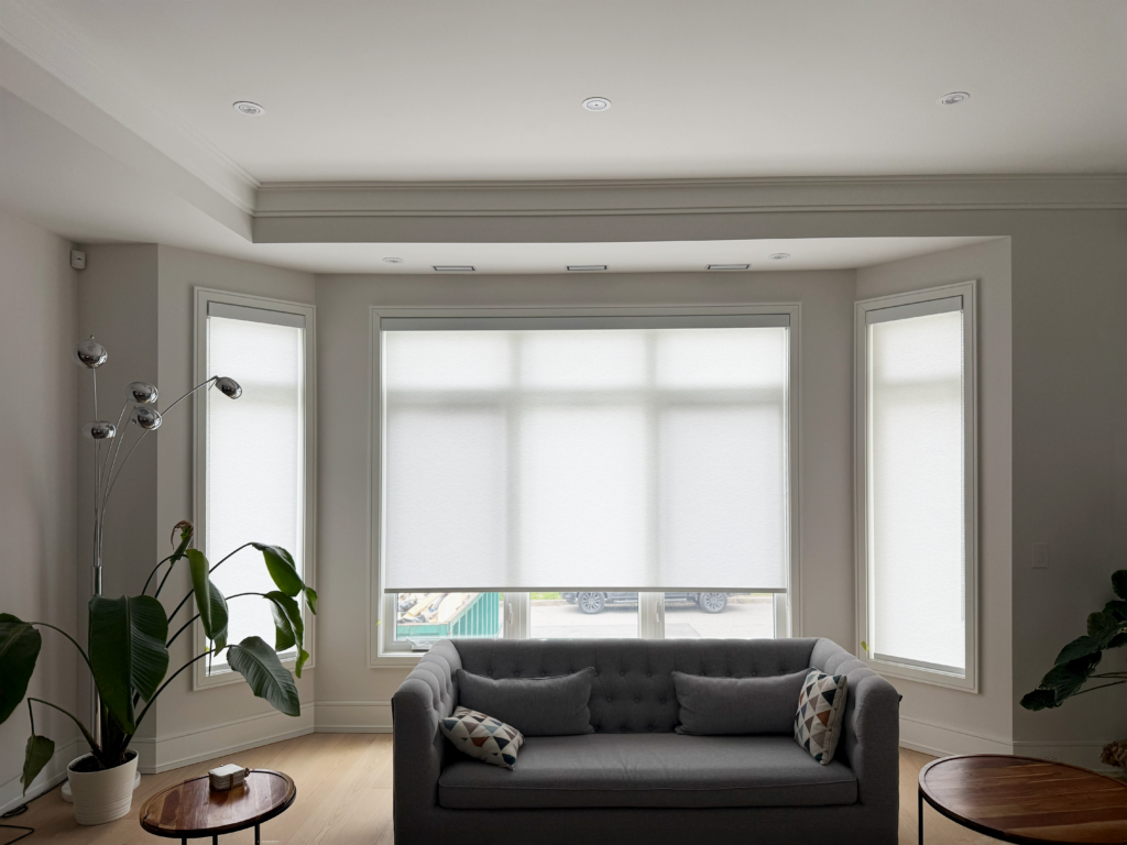 Sleek white custom roller blinds installed on a three-panel bay window behind a grey sofa.