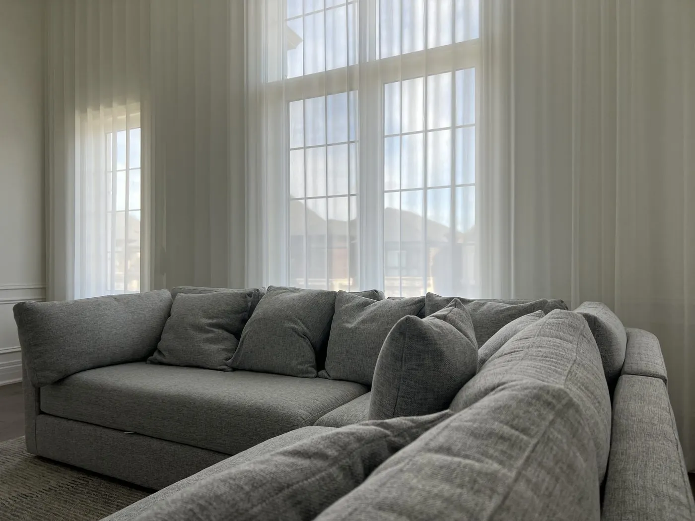 Late-day sun glowing through sheer curtains over two-storey windows behind a grey sectional sofa