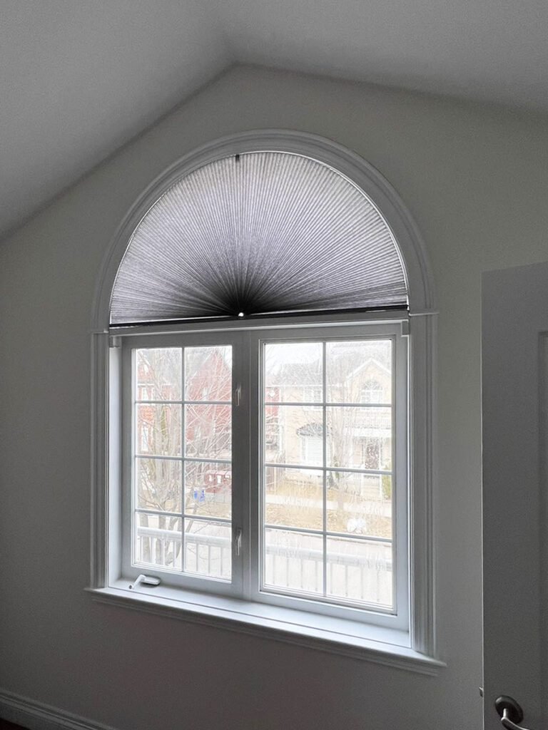 An arched double-hung window in a white-walled room with dark wood flooring, featuring a gray fan-style cellular shade on the top arch looking out onto a residential neighborhood.