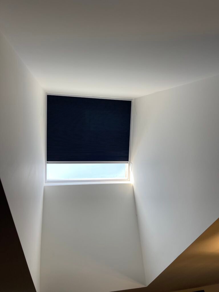 A low-angle view looking up at a deep recessed ceiling skylight equipped with a partially drawn dark blue cellular blackout shade.