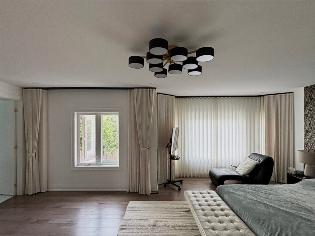 A large bedroom featuring a wide bay window covered with full-length translucent white sheer drapes. Off-white pleated drapery panels are tied back neatly on either side of an adjacent single window, framed by dark ceiling tracks.